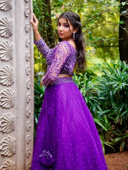 A gorgeous shot from a photoshoot in a garden setting. The model is wearing a vibrant purple lehenga with intricate, full-sleeve beadwork.
