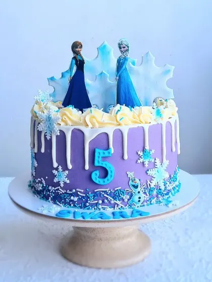 A Frozen-themed drip cake with a purple buttercream base and white chocolate drip. The cake is decorated with edible snowflakes, sprinkles, and character toppers.