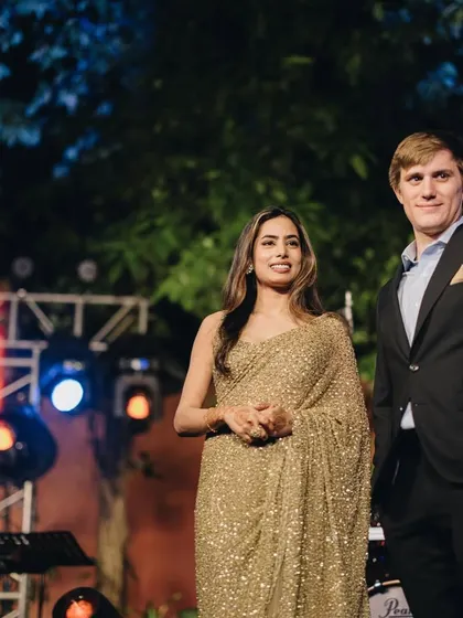 On stage at their cocktail event, the bride's gold saree glitters under the lights, perfectly complemented by the groom's classic black suit.