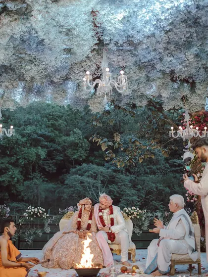 A couple shares a smile during their ceremony, seated under a magical ceiling of white flowers that feels like a cloud. We designed this space to feel like an enchanted forest, creating a warm and beautiful setting for their union.