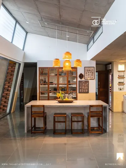The dining island under the high, angled ceiling. The clerestory windows flood the space with natural light, highlighting the simple forms and natural textures of the concrete counter and wooden stools.