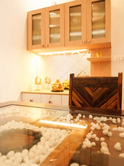 A unique dining table with a glass top and a central portion filled with white pebbles. The custom crockery unit in the background features fluted glass doors and warm lighting.
