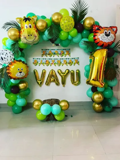 A simple and charming jungle theme balloon arch for a first birthday at home. The setup includes gold letter balloons for the name "VAYU" and cute animal foil balloons.