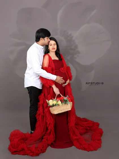 A quiet, intimate moment. The couple embraces, with the mom-to-be holding a basket of flowers, adding a soft and romantic element to this portrait in her red gown.