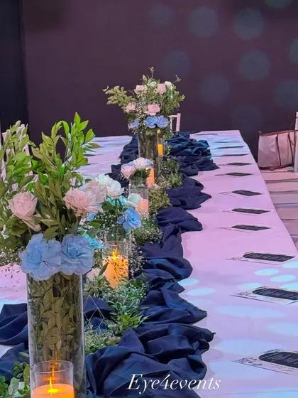 Here is a view of a long dining table decorated in a classic blue and white theme. Tall glass vases with blue and white floral arrangements run down the center, complemented by a navy blue fabric runner and the warm light of candles.