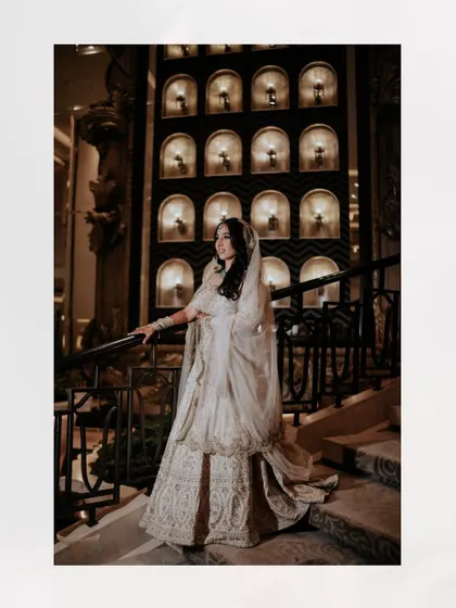 A grand, full-length portrait on a magnificent staircase. The opulent hotel lobby, with its chandelier and intricate railings, provides a stunning backdrop for the bride's elegant white lehenga.