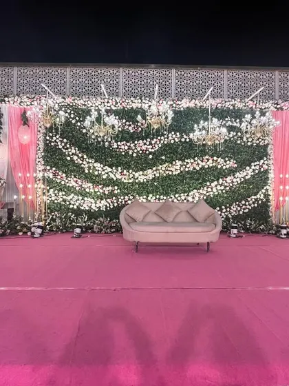 A beautiful outdoor reception stage with a vibrant pink carpet. The backdrop is a lush wall of green and white florals, accented with pink drapes and hanging globe lights.