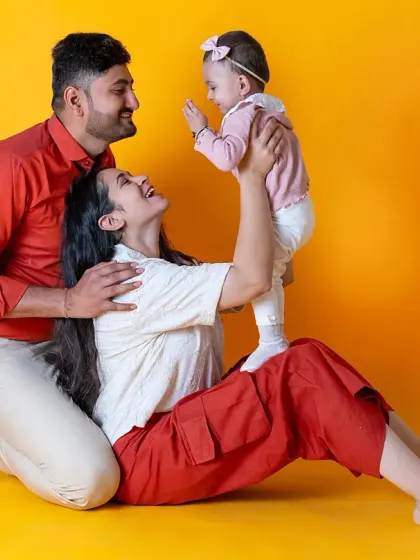 A fun and vibrant family photo against a bright yellow backdrop. The playful interaction between the parents and their baby girl makes for a joyful and energetic portrait.