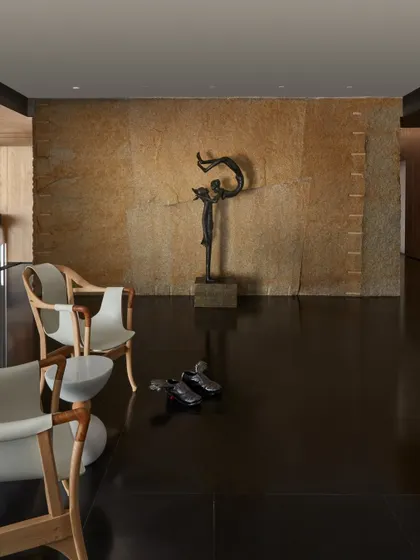 A wide view of the entrance passage, where designer chairs and sculptures are arranged to create a curated, gallery-like feel. The interplay of the rough stone wall, polished black granite floor, and refined art pieces defines our aesthetic.