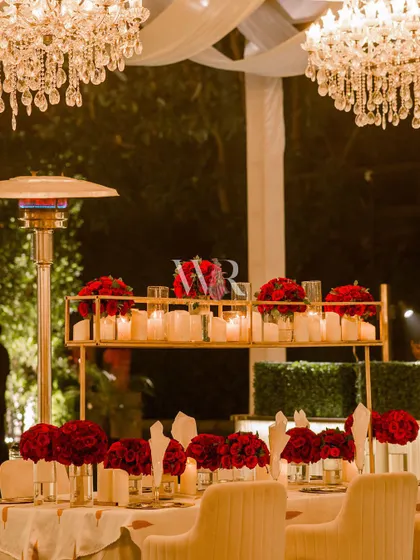 A dining table centerpiece featuring a tiered candle stand and bouquets of deep red roses. The warm candlelight under the glow of chandeliers creates a deeply romantic and intimate dining experience.