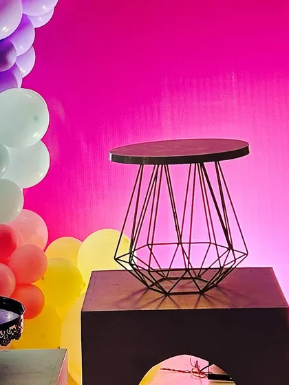 A modern geometric wire table stands on a pedestal, set against a bright pink wall and a rainbow balloon garland.