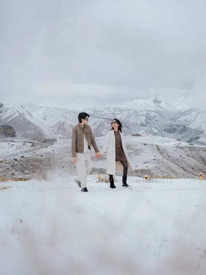 A wide shot capturing the vast, snow-covered mountains of Azerbaijan. The couple walks hand-in-hand, showcasing the grand scale and epic nature of a destination pre-wedding shoot.
