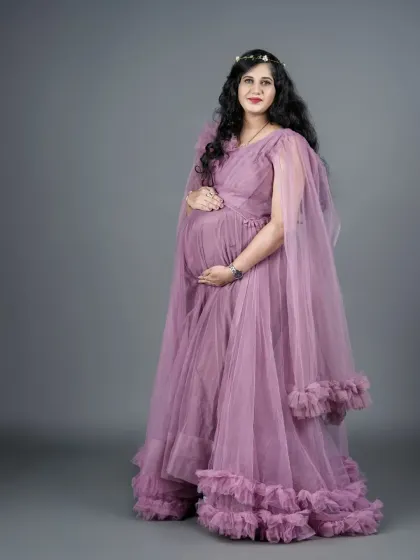 A full-length portrait of an expecting mother in a light purple, floor-length gown with ruffled details. The simple grey studio background puts all the focus on her serene expression and beautiful baby bump.