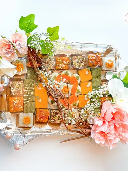 This hamper showcases a diverse assortment of gourmet mithai on an elegant silver-plated tray. I love combining different colors, textures, and flavors, all brought together with a delicate floral and twig arrangement.