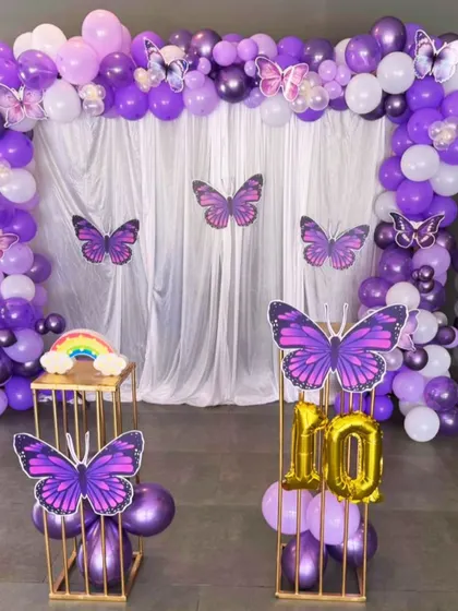 A magical butterfly theme for a 10th birthday, using shades of purple and white balloons. The setup includes a full balloon arch over a white curtain backdrop, with beautiful purple butterfly cutouts appearing to flutter around the scene.