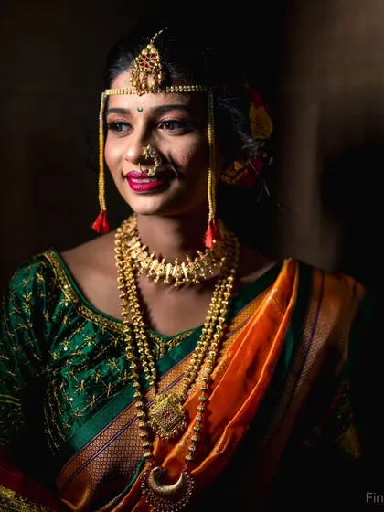 A dramatic, low-key portrait of a Maharashtrian bride. The lighting sculpts her features and makes the gold jewelry stand out against the dark background.