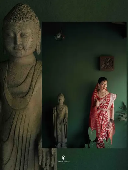An artistic portrait of the bride in her red saree, standing next to a Buddha statue, blending tradition with tranquility.