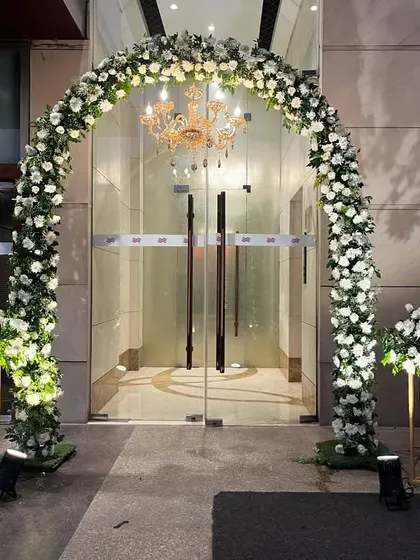 A grand floral arch at the entrance of the Crowne Plaza for a Nikaah ceremony. The arch is covered in white roses and greenery, with a classic chandelier hanging in the center.