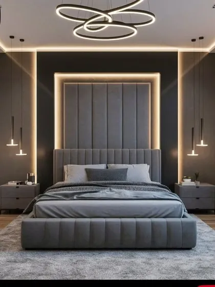 This master bedroom design creates a dramatic and luxurious feel with a floor-to-ceiling upholstered headboard, highlighted by recessed LED backlighting. The layered lighting scheme, including a modern chandelier and hanging bedside pendants, adds depth and sophistication to the contemporary grey palette.