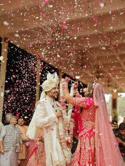 A joyful and dynamic wedding moment capturing the bride and groom during the Varmala ceremony amidst a shower of flower petals. This candid shot freezes a moment of pure celebration and happiness.