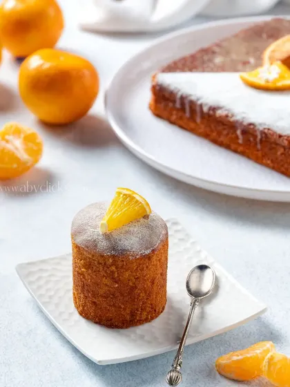 A mini orange drizzle cake with a dusting of powdered sugar, styled with fresh oranges for a bright and zesty product shot.