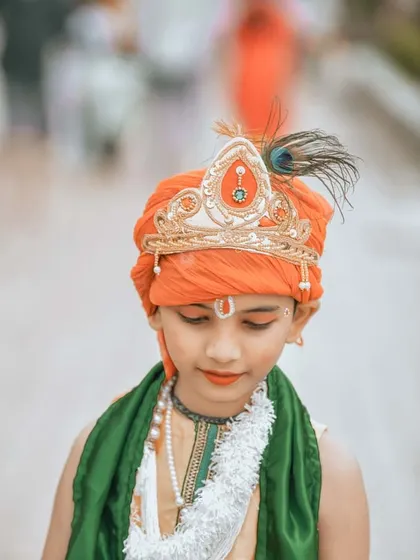 A close-up portrait of the young boy dressed as Krishna. This shot focuses on the details of his orange turban with a peacock feather and his serene, contemplative expression, highlighting the cultural richness of the moment.