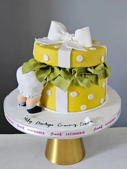 A playful and adorable baby shower cake designed to look like a gift box. The cake features a cute baby bottom peeking out, a big white bow, and a cheerful yellow polka dot pattern.