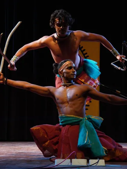 A powerful pose from a Mayurbhanj Chhau performance, highlighting the use of traditional weaponry. The dancer in front wields a curved sword, while the one behind holds a stringed instrument, showing the blend of combat and rhythm.