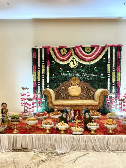 A wide shot of a Seemantham stage with a greenery wall and floral arch. The setup includes a welcome easel and a full display of traditional offerings in front of the ornate sofa.