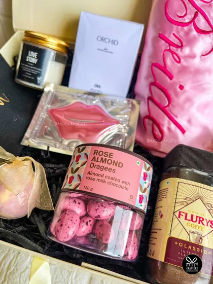 A close-up of a bride-to-be hamper, showcasing a 'Love Story' candle, Zara perfume, a lip mask, and rose almond dragees.