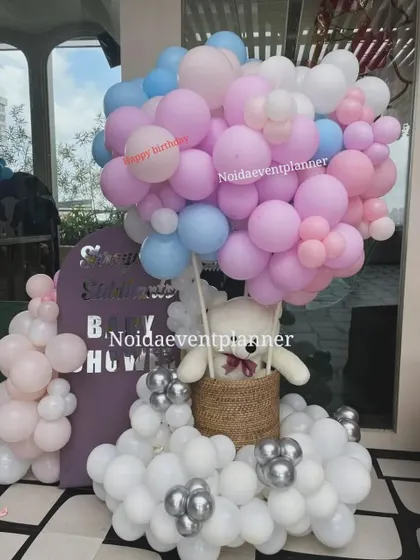 A hot air balloon concept for a baby shower, using a basket with a teddy bear and a large cloud of pink, blue, and white balloons.