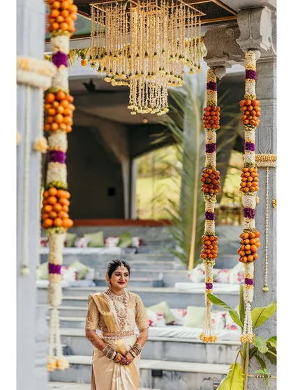 A beautiful bride stands framed by traditional floral decor. The hanging arrangements of oranges and flowers, along with a floral chandelier, create a unique and festive look for her ceremony.