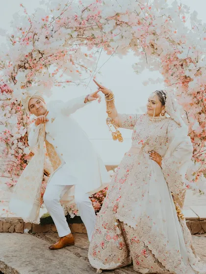 A playful and quirky pose in front of a pink floral arch. This image shows the fun personality of the couple, adding a touch of lightheartedness to their elegant wedding album.
