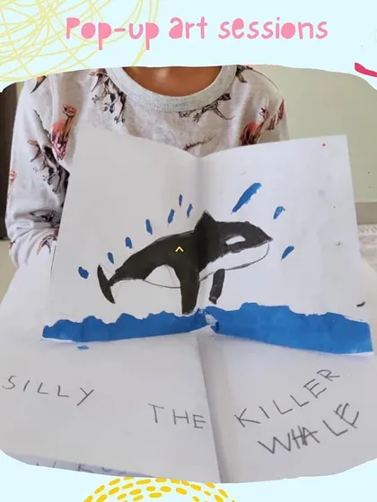A child proudly displays a pop-up book they created featuring a killer whale. Our pop-up sessions teach basic paper engineering skills to bring stories to life in 3D.