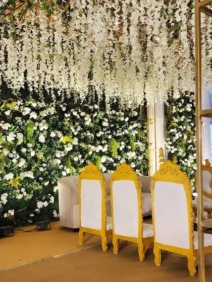 A lush green and white themed Vidhi setup inside a banquet hall. The stage features a dense floral wall and a ceiling of hanging wisteria, creating an indoor garden effect.