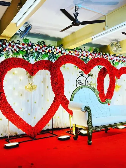 An elegant and romantic reception stage featuring two large, interlocking heart-shaped frames covered in red flowers. A classic white chaise lounge sits in front, set against a white curtain backdrop, perfect for a love-themed wedding.