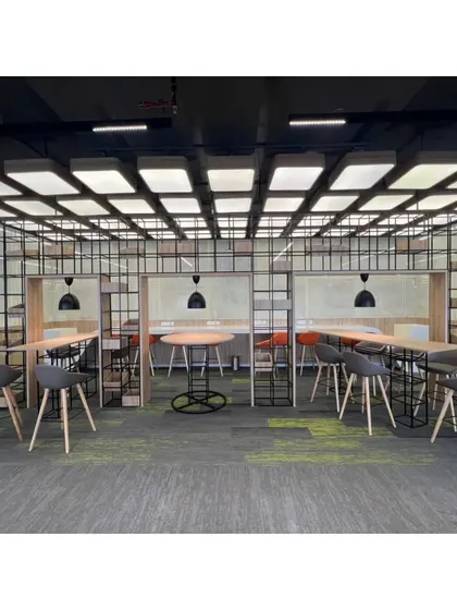 A semi-enclosed collaborative hub within the open office, featuring high tables and stools. The metal grid shelving provides a sense of separation without sacrificing the open feel, making it an ideal space for informal team meetings.