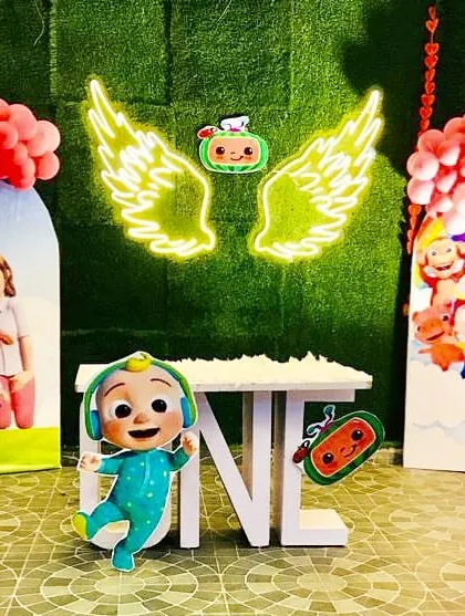 A Cocomelon-themed first birthday with a unique neon angel wings backdrop. The setup includes character cutouts and a "ONE" table.