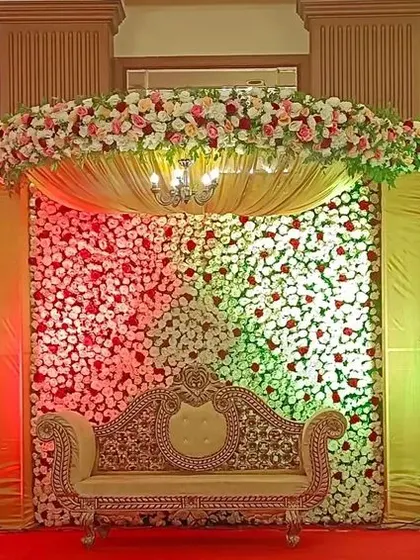 A traditional reception stage with a mix of floral walls and rich golden drapery. The layered look and classic furniture create a grand and festive atmosphere for a wedding celebration.