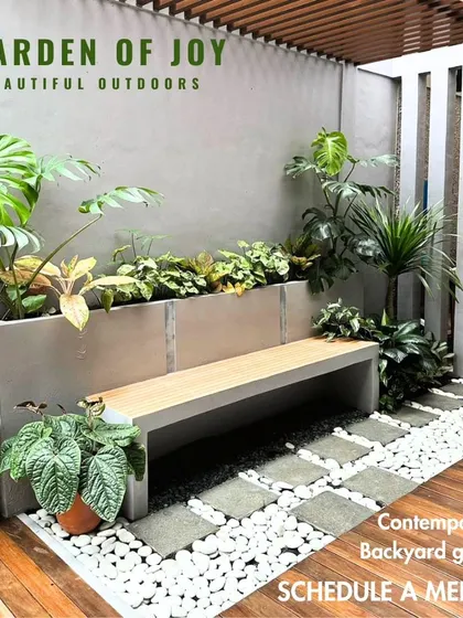 This contemporary backyard design shows how we integrate hardscape and softscape elements. A built-in concrete planter with a bench is surrounded by lush monstera plants, with a pathway of stone pavers and white pebbles on a wooden deck.