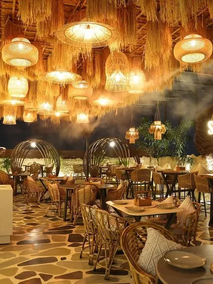 The interior of Sesh Delhi, designed to capture the vibrant, bohemian spirit of Goa. The ceiling is a dense tapestry of straw and woven pendant lights, creating a dramatic, immersive experience complemented by natural stone flooring and rattan furniture.
