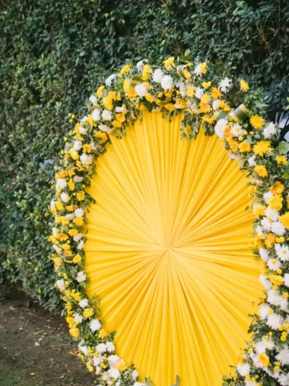 A bright and sunny photo backdrop for a Haldi ceremony, featuring a circle of pleated yellow fabric framed with yellow and white chrysanthemums.