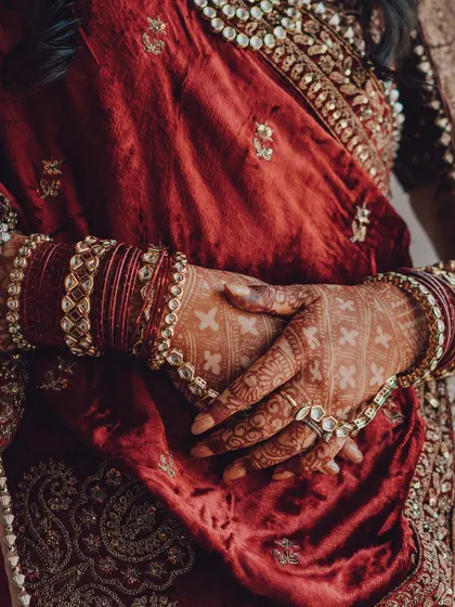 A detailed shot of a bride's hands, adorned with intricate henna and traditional bangles, resting on a rich velvet outfit. We believe the small details tell a big part of the wedding story.
