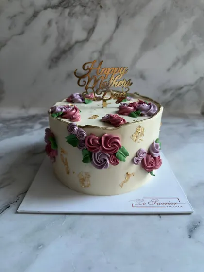 A beautiful Mother's Day cake featuring hand-piped buttercream roses in shades of purple and pink, accented with green leaves and flecks of edible gold leaf.
