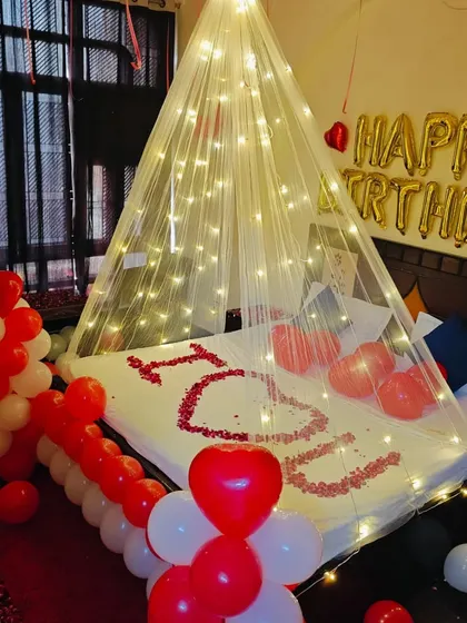 A romantic birthday surprise with a fairy light canopy. The bed is decorated with 'I ❤️ U' in rose petals, surrounded by red and white balloons.