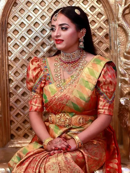 A truly royal bridal look. The bride is seated on a throne-like chair, wearing a multi-colored silk saree with puff sleeves. Her makeup is bold and elegant, with a red lip and diamond jewellery.