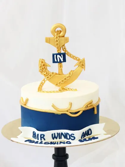 A farewell cake for a retiring Naval Officer, wishing them "Fair winds and following seas." The design features a striking gold fondant anchor and chain on a navy blue and white base, symbolizing a distinguished career.