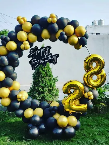 An outdoor 28th birthday setup with a black and gold balloon ring and large foil number balloons.