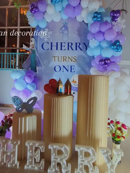 A 'Frozen' themed first birthday party for 'Cherry'. The setup features a character backdrop with Elsa and Olaf, a balloon arch in icy blue, purple, and white, and snowflake details.