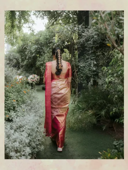 A stunning back shot of the South Indian bride, showcasing her traditional braided hairstyle with intricate gold ornaments as she walks through a garden path.